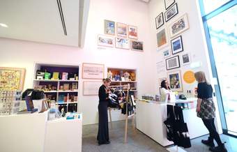 A photograph of Tate Liverpool's shop at RIBA North. One visitor is looking through some bags on display and another visitor is being served at the till by a member of staff.