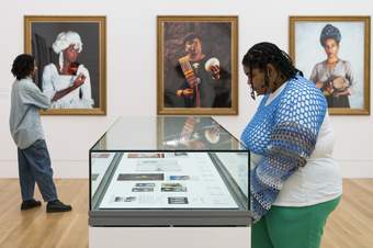 One visitor browses three large photographic portraits in wooden frames on the wall. One visitor peers into a glass display case in the centre of the room containing several small prints.