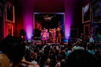 Three performers wearing extravagant outfits on a stage in an art gallery, lit by purple and pink lighting