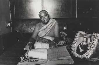 A figure sits on the floor in an interior setting, surrounded by papers and a framed photograph of a person graduating.