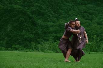 Two dancers in loose brown clothing moving in a green field, with a cluster of green trees in the background