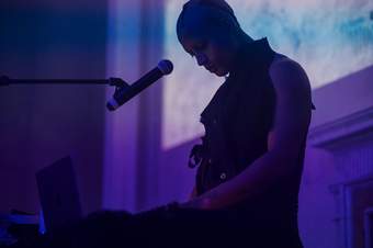 Photograph of Joshua Woolford standing in purple light with a laptop and microphone in front of them