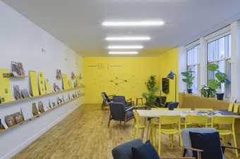 A room with tables and chairs in it. On the left wall are shelves which have printed resources on them.