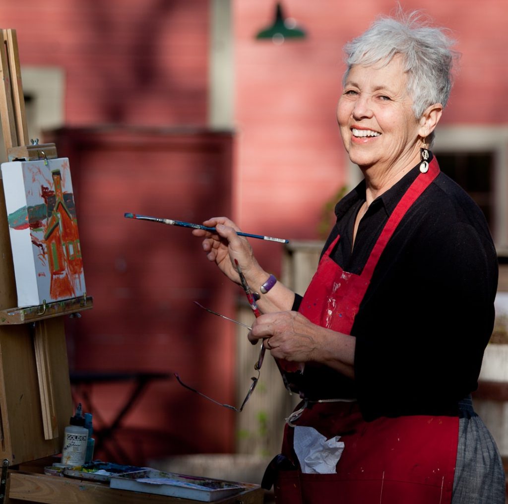 Louise Minks in front of a red mill, holding a paintbrush and smiling as she paints the mill