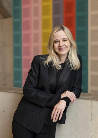 photo of a light skinned woman with light hair in a black jacket standing in front of a wall with vertical stripes