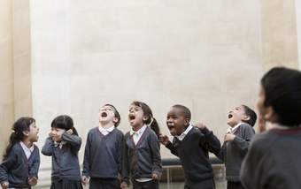 A group of children dressed in school uniform shout out loud. One child covers their ears and smiles.