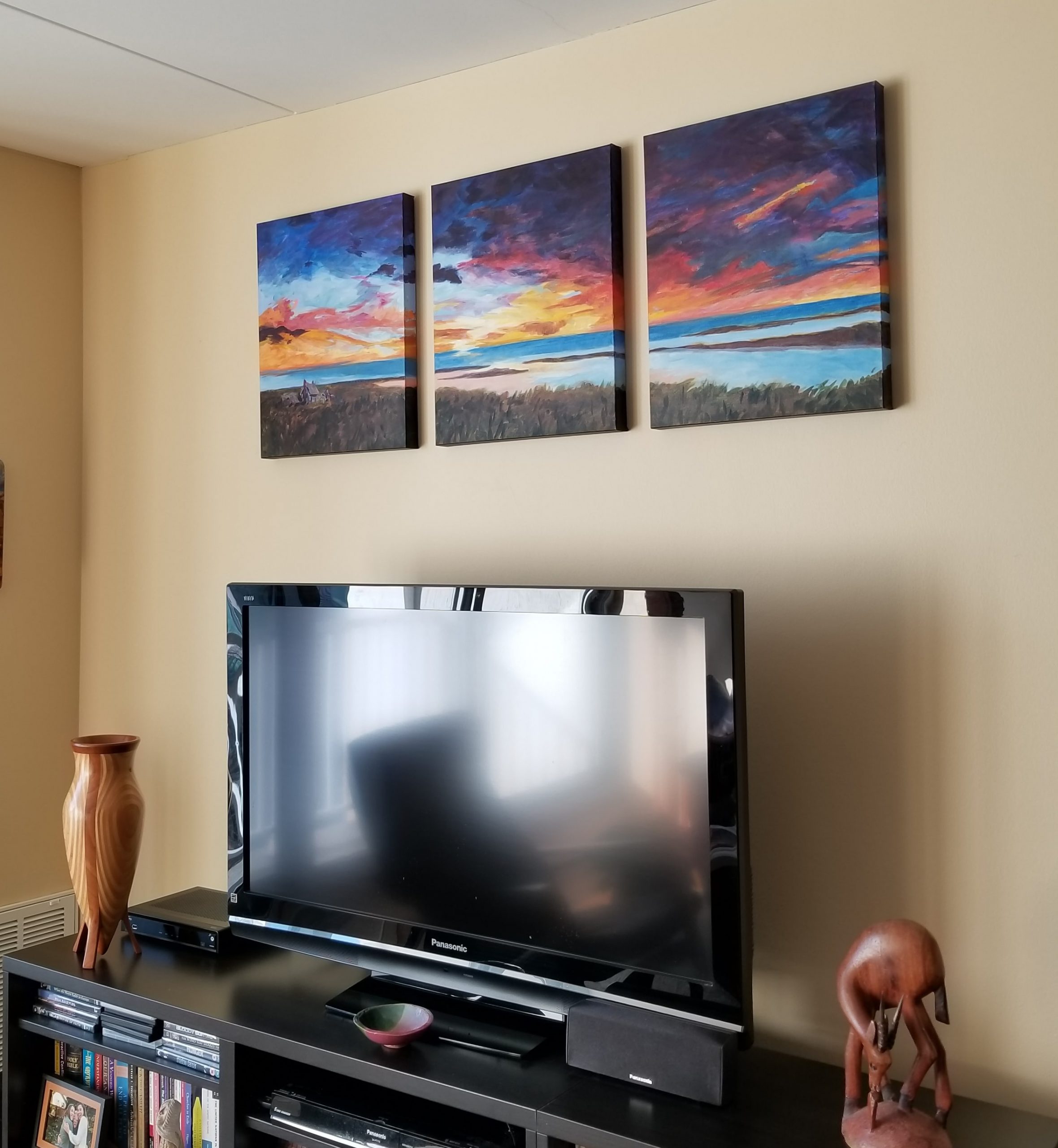 A set of three paintings showing a marshy landscape at sunset over a living room television