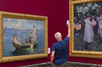 A person taking a photo on a smart phone of a painting in a gold frame on a dark red wall