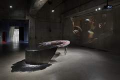 A modern art installation featuring a sculptural table with a colourful surface on one half and a dark grey, liquid-like surface on the other half, situated in a gallery with concrete walls and floor. A projection of figures is displayed on the back wall.